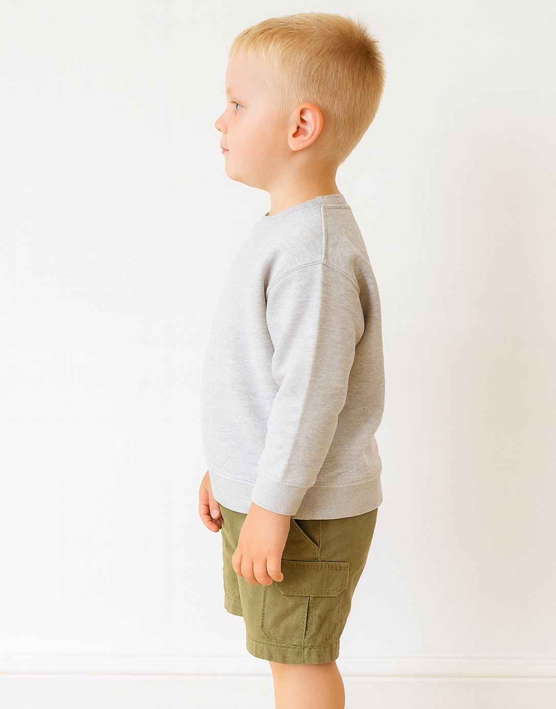 Side view of young boy wearing a gray sweatshirt and green shorts on a white background