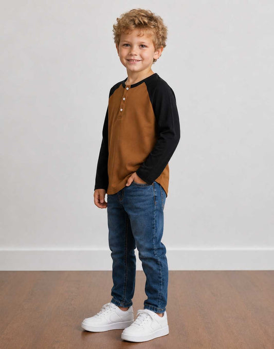 Side view of a young boy with curly blond hair stands in a studio wearing a brown and black Raglan Long Sleeve Henley Top, blue jeans, and white sneakers against a light gray wall and wood floor.