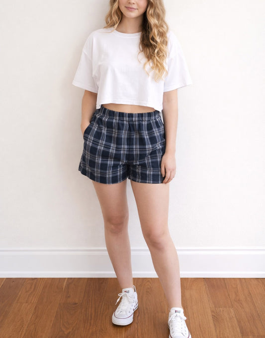 Woman wearing white tee with blue and white plaid Easy Elasticized Waist Shorts and white shoes with wooden floor and plain background.