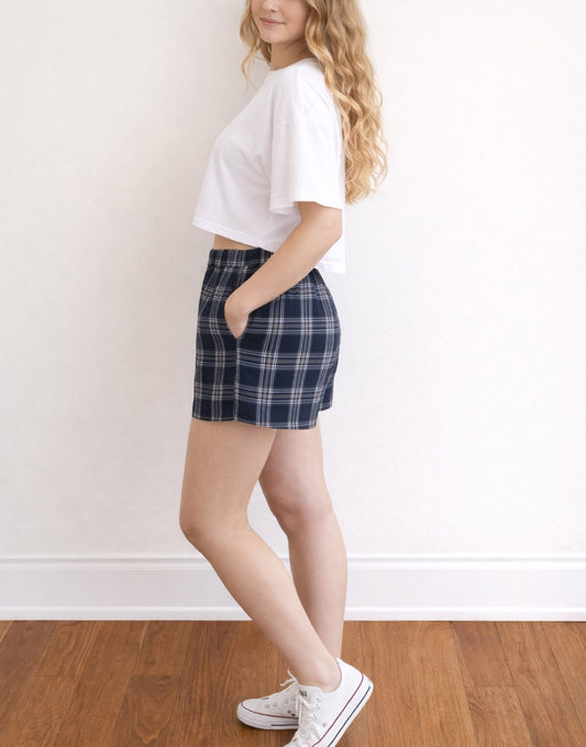 Side view of woman wearing white tee with blue and white plaid Easy Elasticized Waist Shorts and white shoes with wooden floor and plain background.