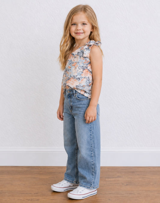 Side view of a young girl with blonde hair stands against a white textured wall wearing a sleeveless floral One Shoulder Top with a tie detail, wide-leg blue jeans, and white sneakers.