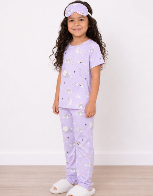 Side view of a young girl stands against a white wall wearing a light purple Pajama Set with a moon and star print, a matching sleep mask, and white slippers.