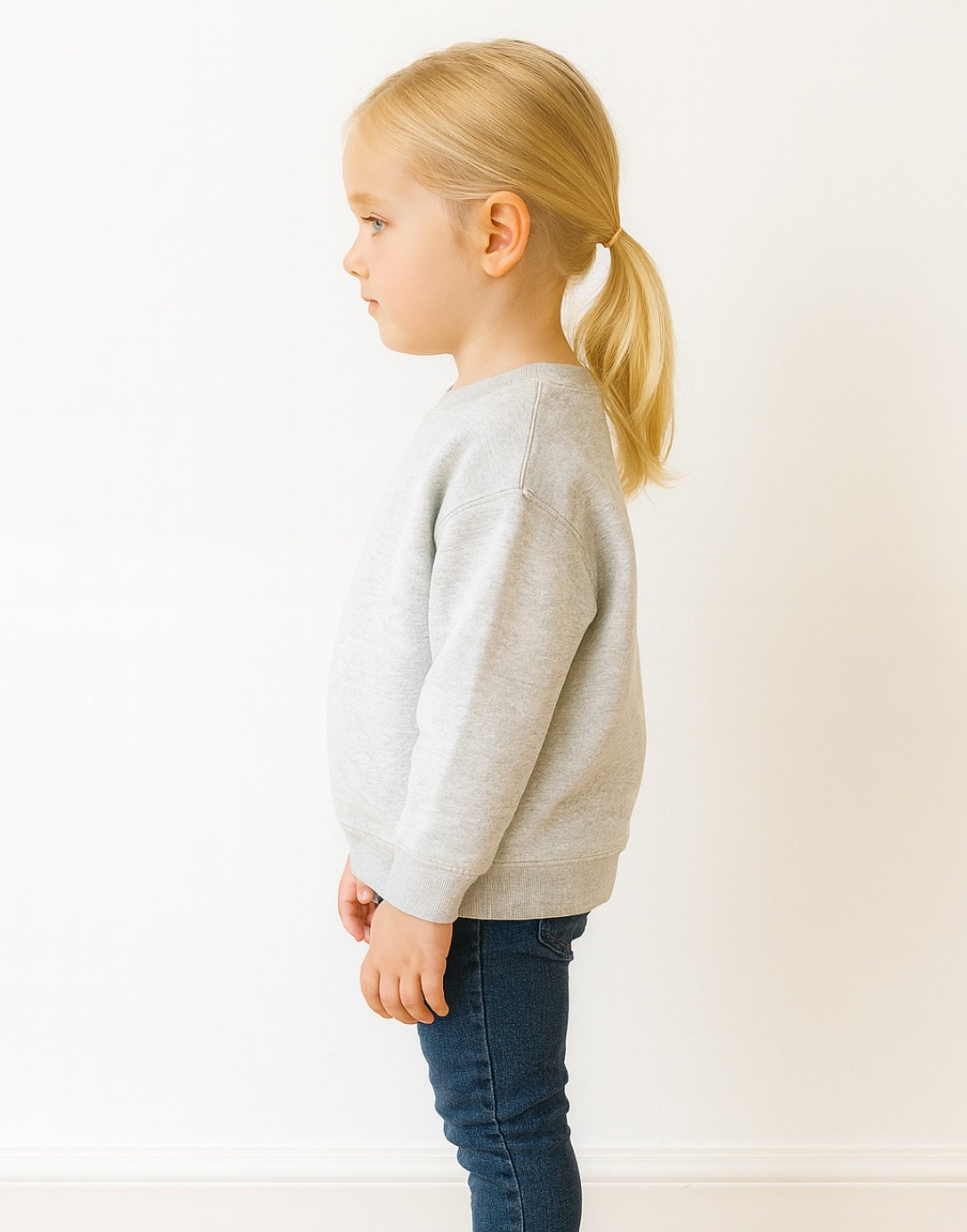 Side view of young girl wearing a gray sweater and blue jeans standing against a white background