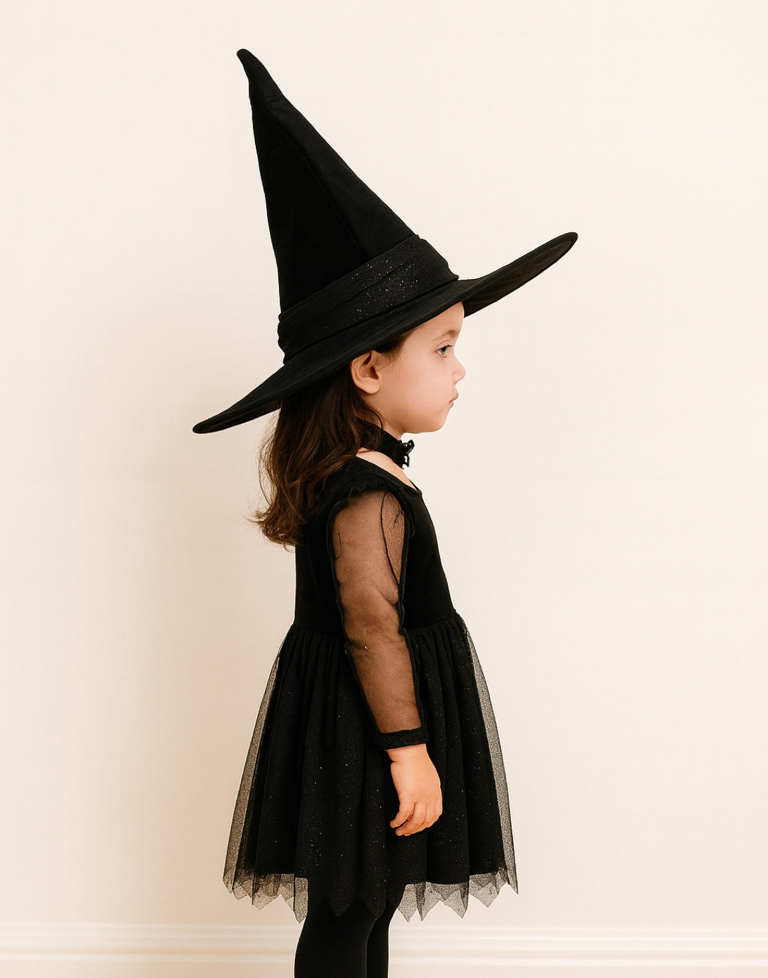 Side view of child wearing a black witch costume with a pointed Witch Hat on a plain background.