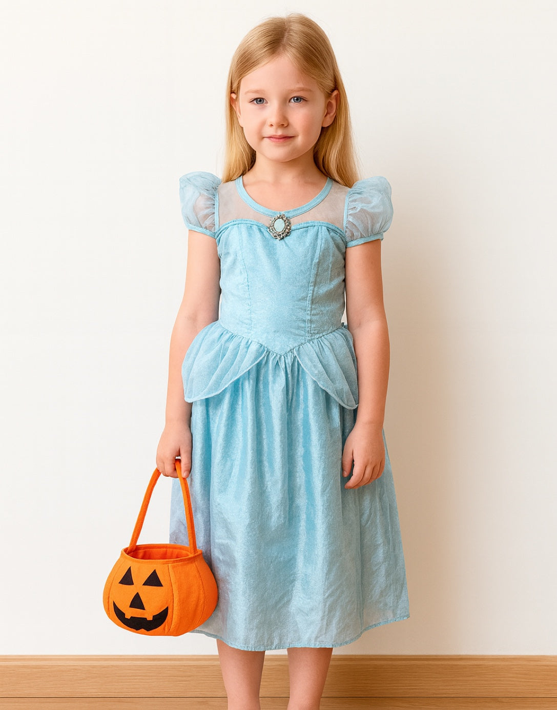 Young girl in a light blue dress holding an orange Halloween Pumpkin Trick Or Treat Bag against a plain background.
