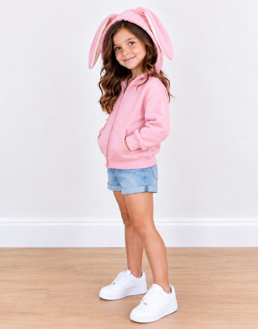 Side view of a young girl wearing a pink hooded sweatshirt with long bunny ears, light denim shorts and white shoes on a plain background.
