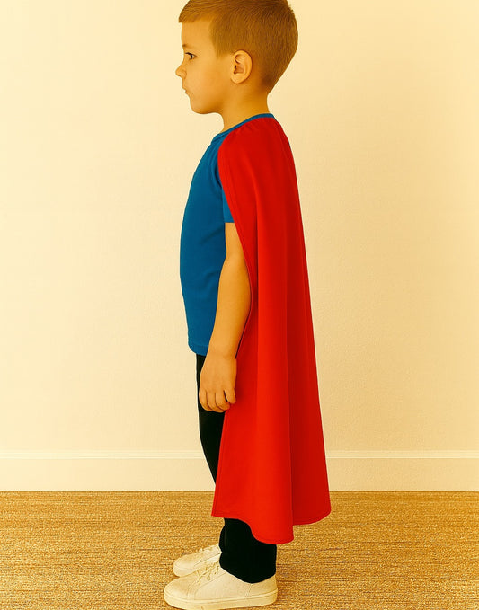 Side view of kid wearing a red cape over a blue shirt, and black pants on a beige background.