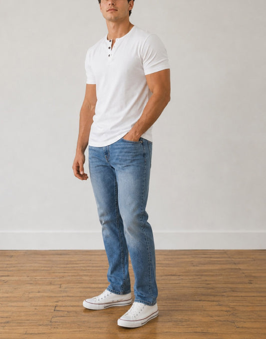 Side view of a man wearing a white Short-Sleeve Henley Tee Shirt, blue jeans, and white canvas sneakers, standing in a room with a light gray wall and wooden floor.
