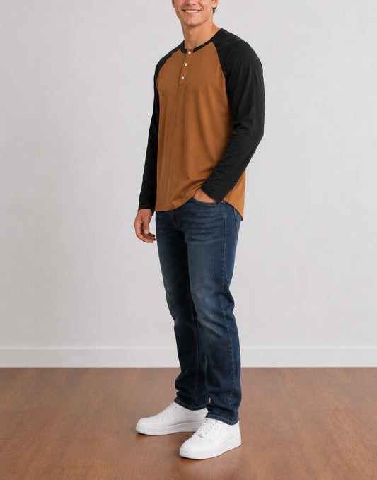 Side view of a man wearing a brown and black Raglan Long Sleeve Henley Top, dark blue jeans, and white sneakers, standing against a light gray wall and wood floor.