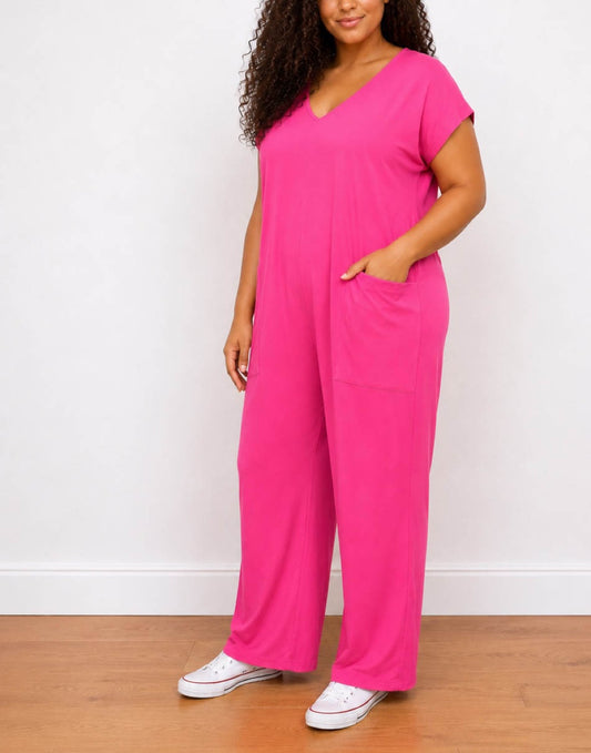 Side view of a woman wearing a bright pink short-sleeve Plus Size Oversized Knit Tee Jumpsuit with a V-neck and side pockets, paired with white sneakers, standing against a white wall and wooden floor.