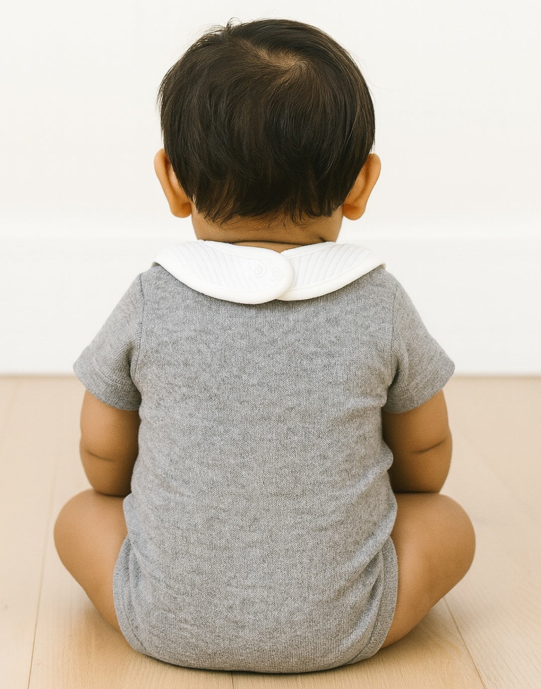 Back view of baby wearing a gray onesie with a white collar sitting on a light-colored surface.