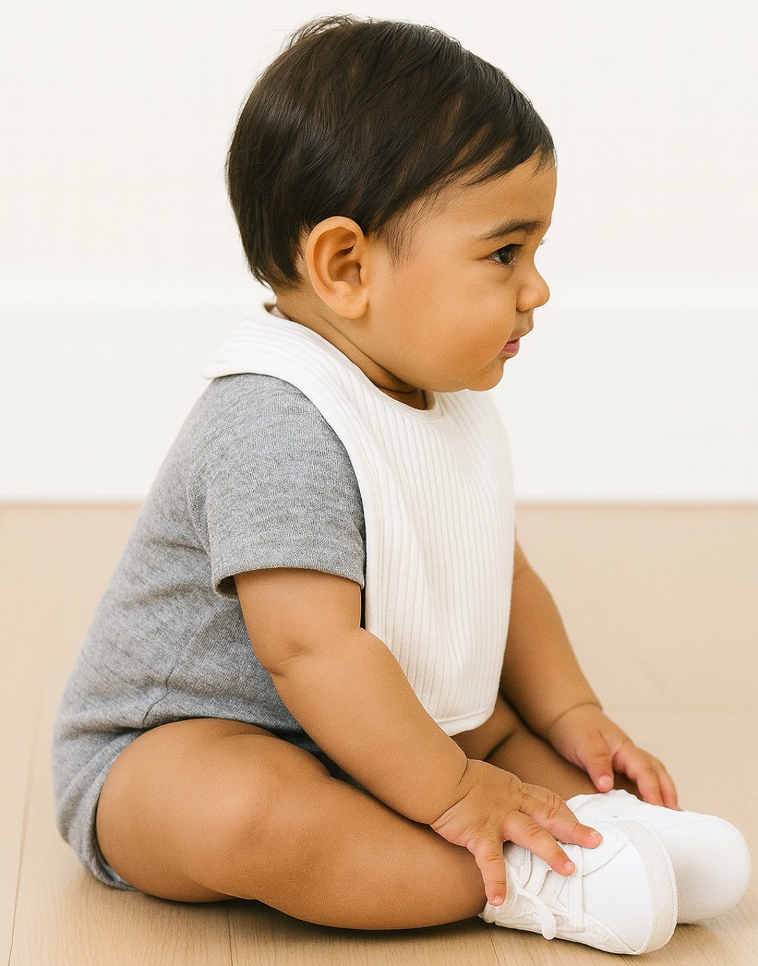 Side view of baby sitting on a light-colored floor wearing a gray shirt, white bib, and white shoes.