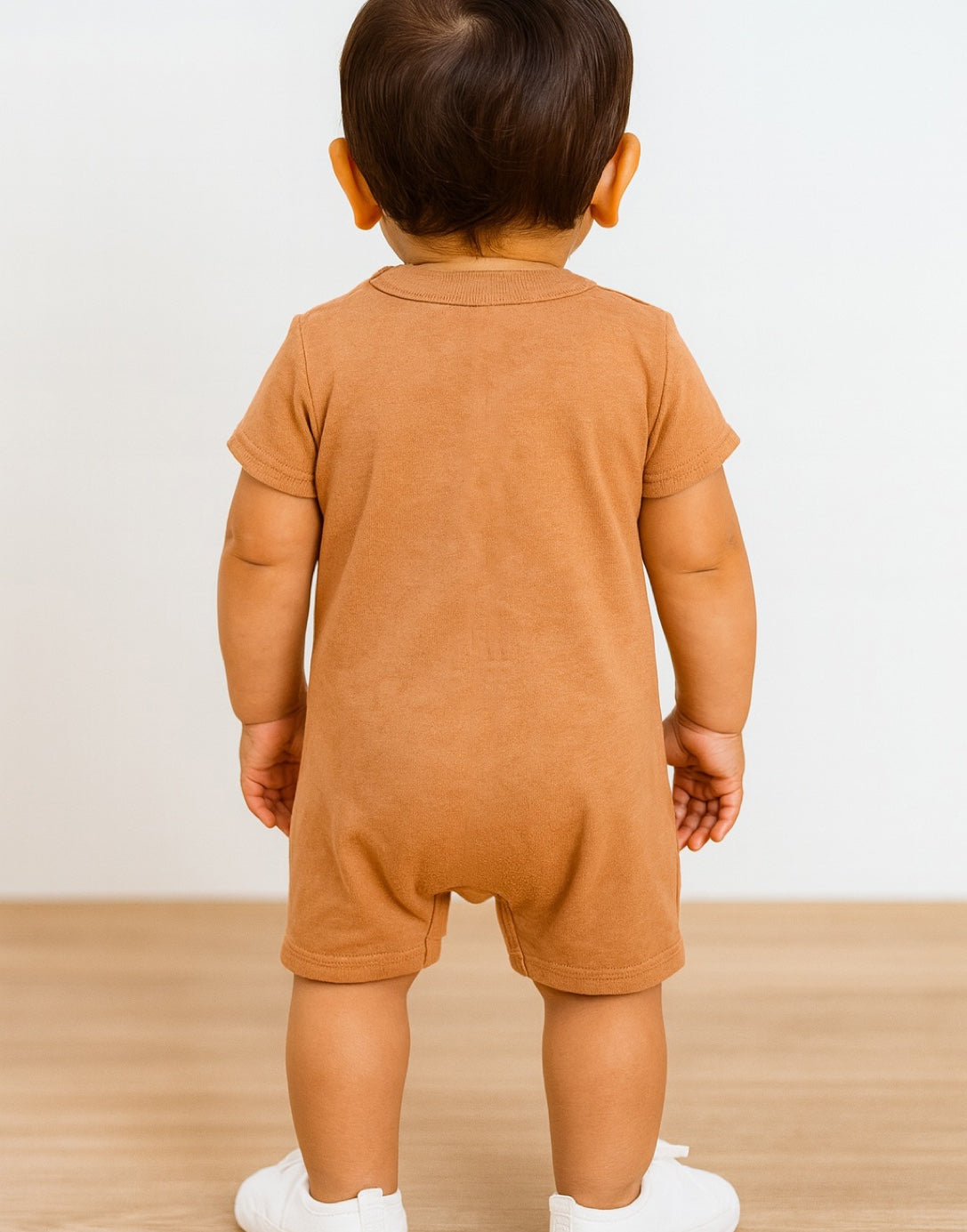 Back view of baby wearing a brown Crew Neck T-Shirt Romper onesie on a light wooden floor with a white wall background