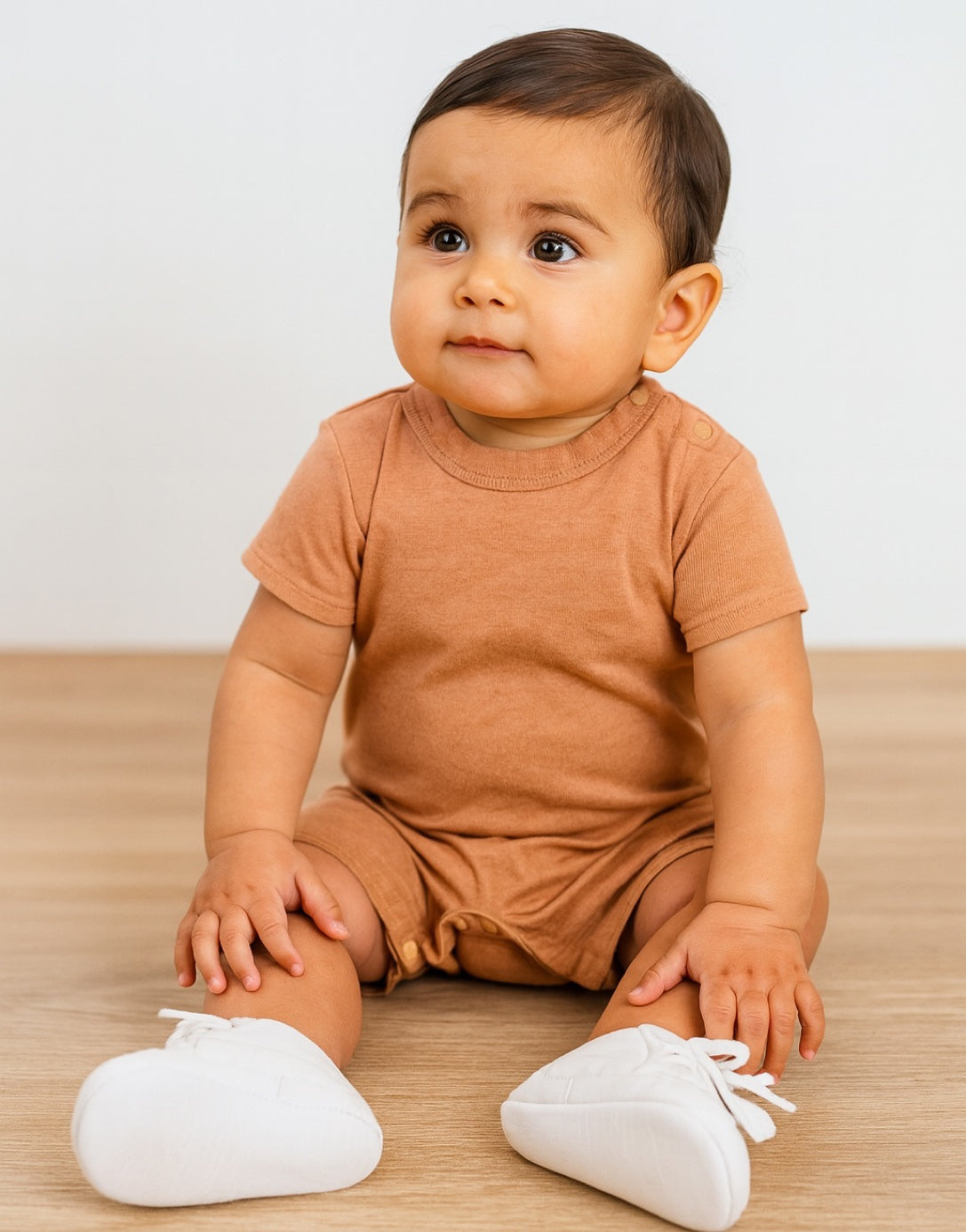 Baby wearing a brown Crew Neck T-Shirt Romper with white shoes on a wooden floor.