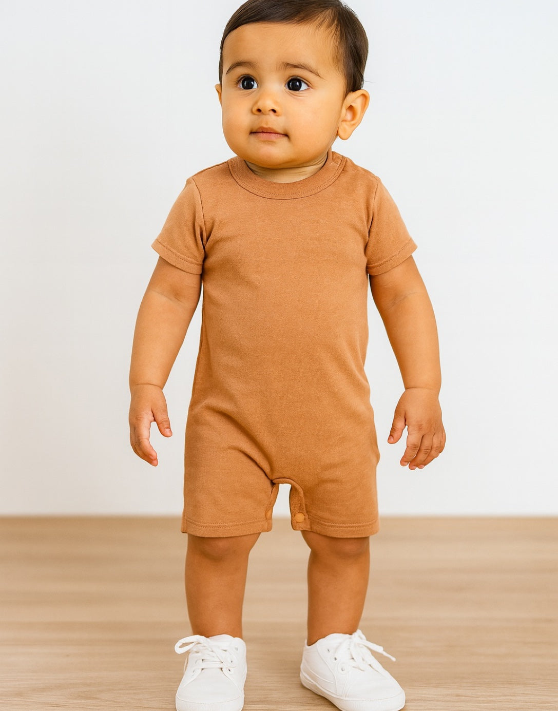 Side view of baby wearing a brown Crew Neck T-Shirt Romper onesie with white shoes on a wooden floor.