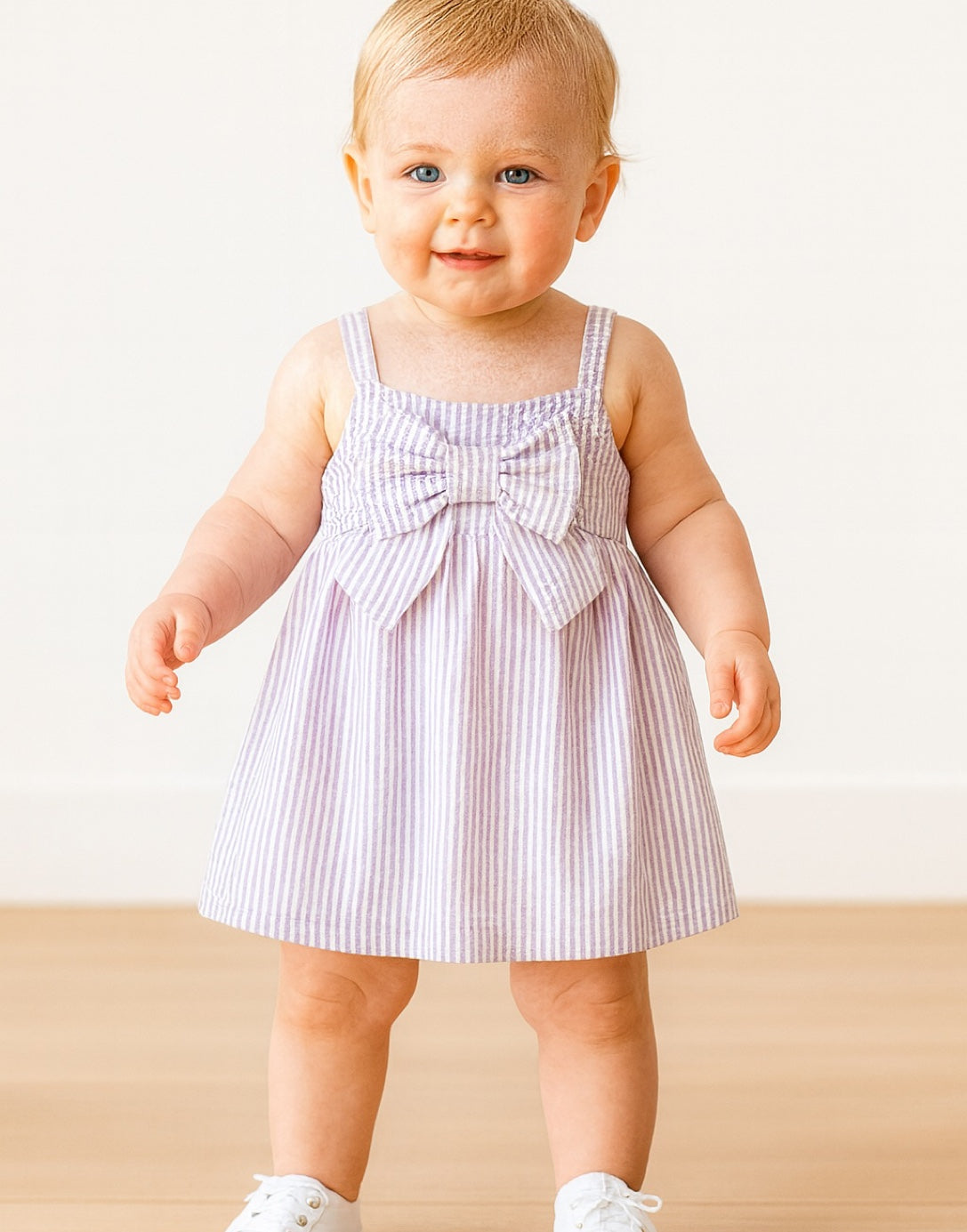 Baby wearing a striped Baby Girl Bow Front Cami Dress with a bow on a beige background