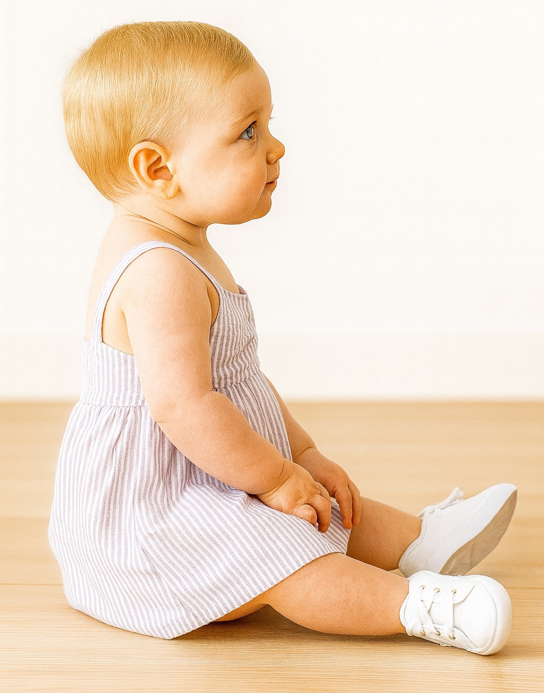 Side view of baby sitting on a wooden floor wearing a striped Baby Girl Bow Front Cami Dress and white shoes.