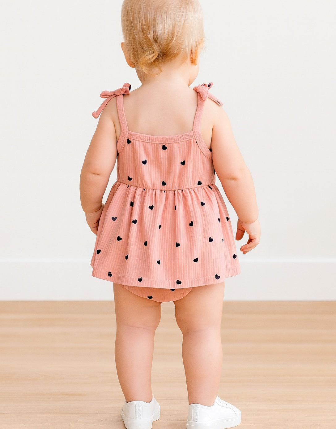 Back view of child wearing a pink Baby Girl Cami Bodysuit Dress with black heart patterns on a light wooden floor.
