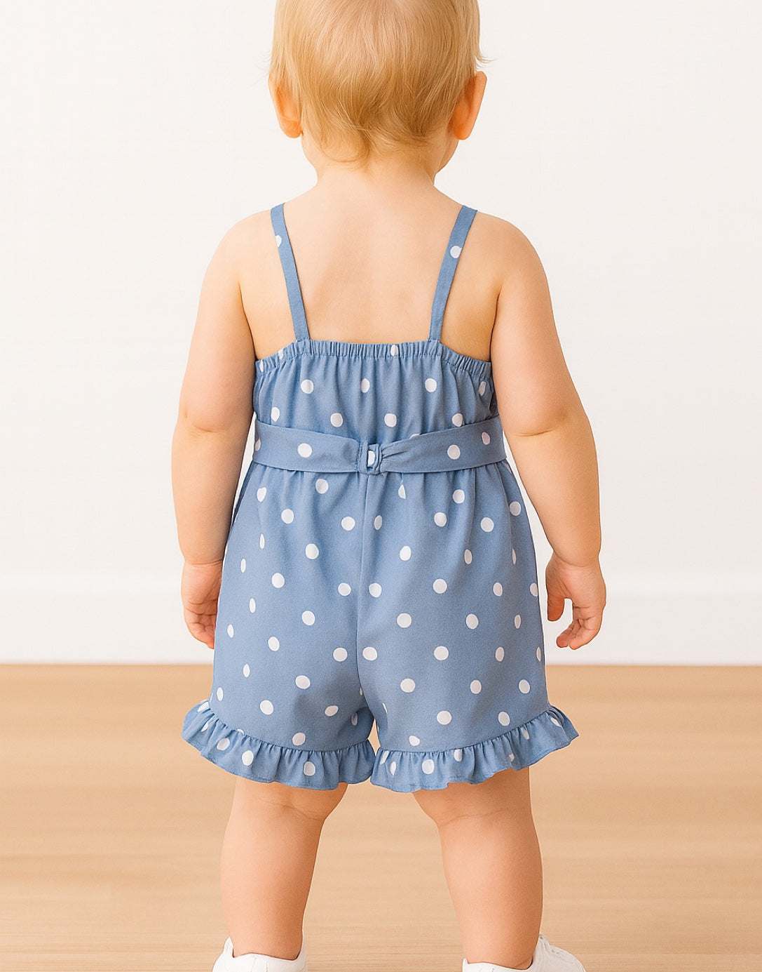 Back view of baby wearing a blue polka dot Cami Ruffle Hem Romper on a light background