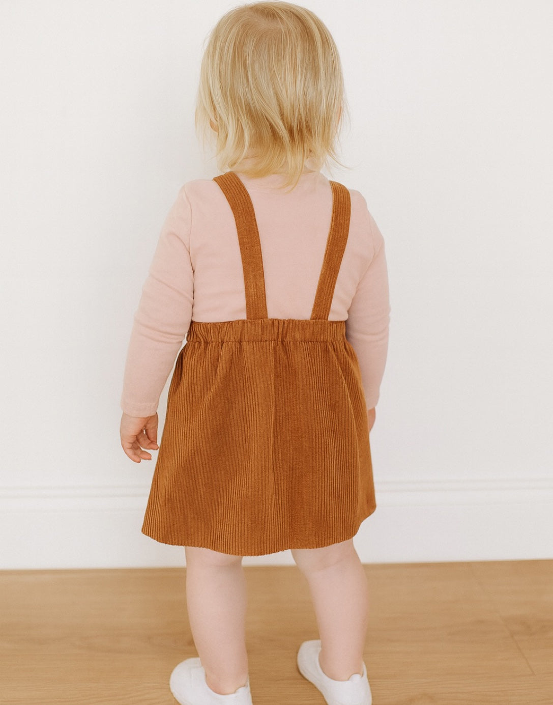 Back view of child wearing a brown Overall Dress and white shoes in a softly blurred indoor setting