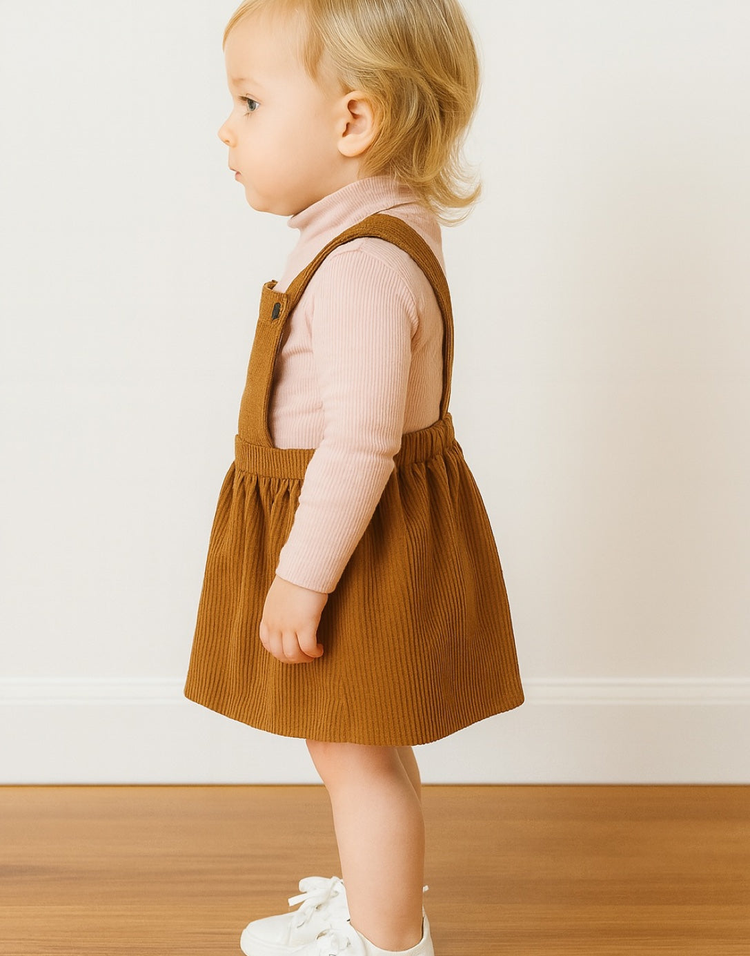 Side view of child wearing a brown Overall Dress with a pink shirt against a white wall.