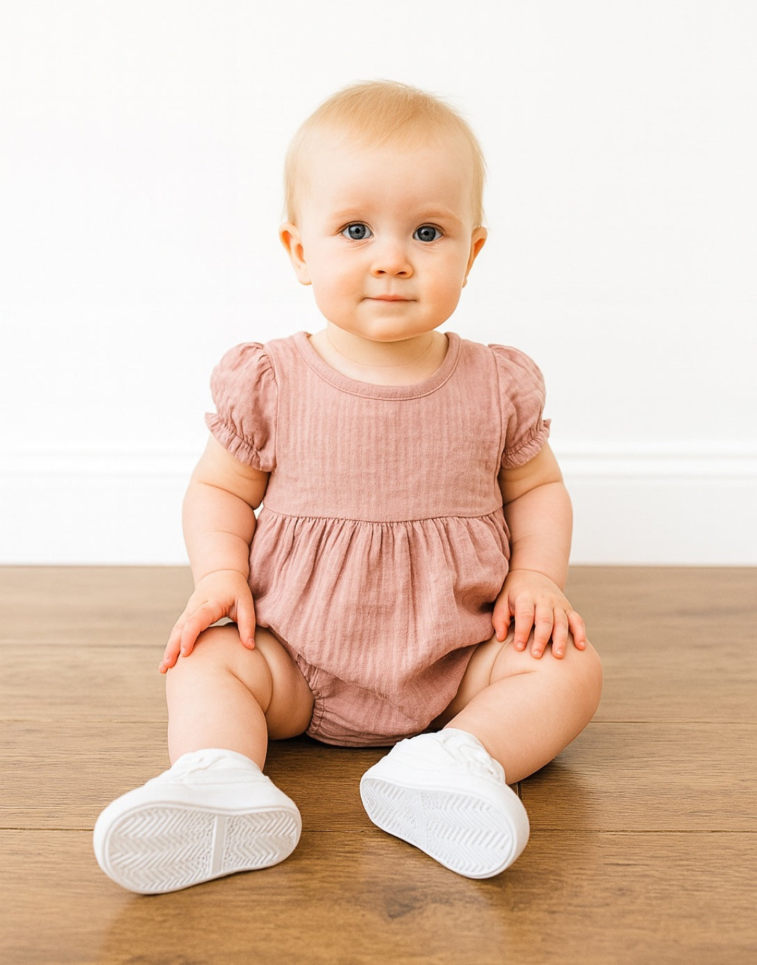 Baby sitting on a wooden floor wearing a pink Puff Sleeve Onesie Romper and white shoes.