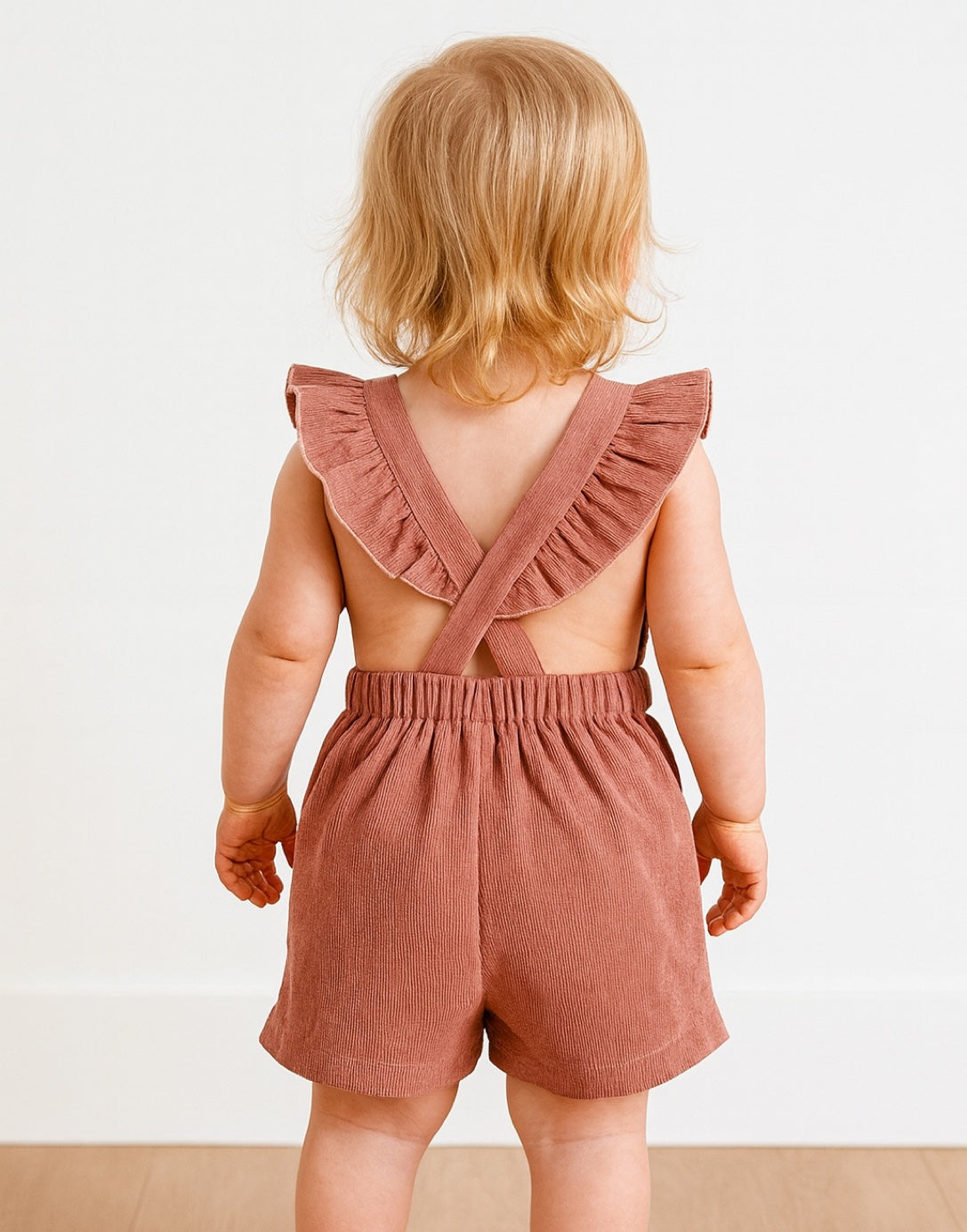 Back view of child wearing a pink romper with ruffled straps on a white background