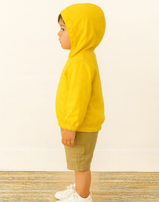 Side view of child wearing a yellow Windbreaker Jacket and beige shorts on a wooden floor with a light-colored wall background