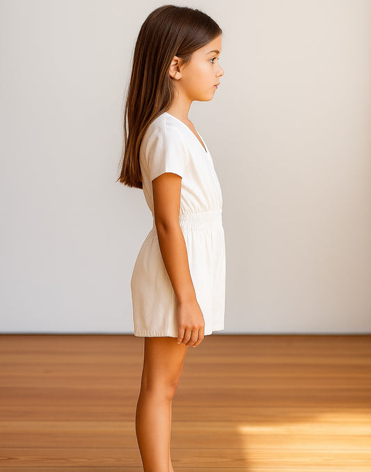 Side view of young girl wearing a white Kimono Sleeve Surplice Romper against a plain background.