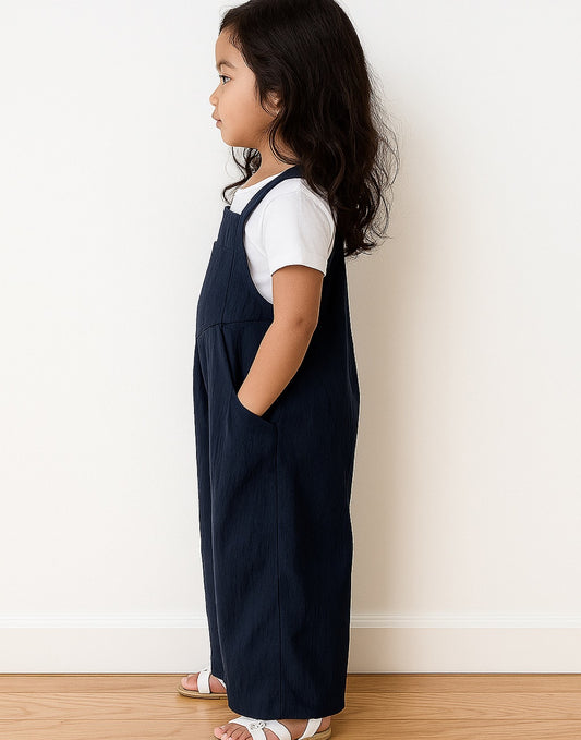 Side view of young girl wearing navy blue Pinafore Jumpsuit and a white shirt against a white wall.