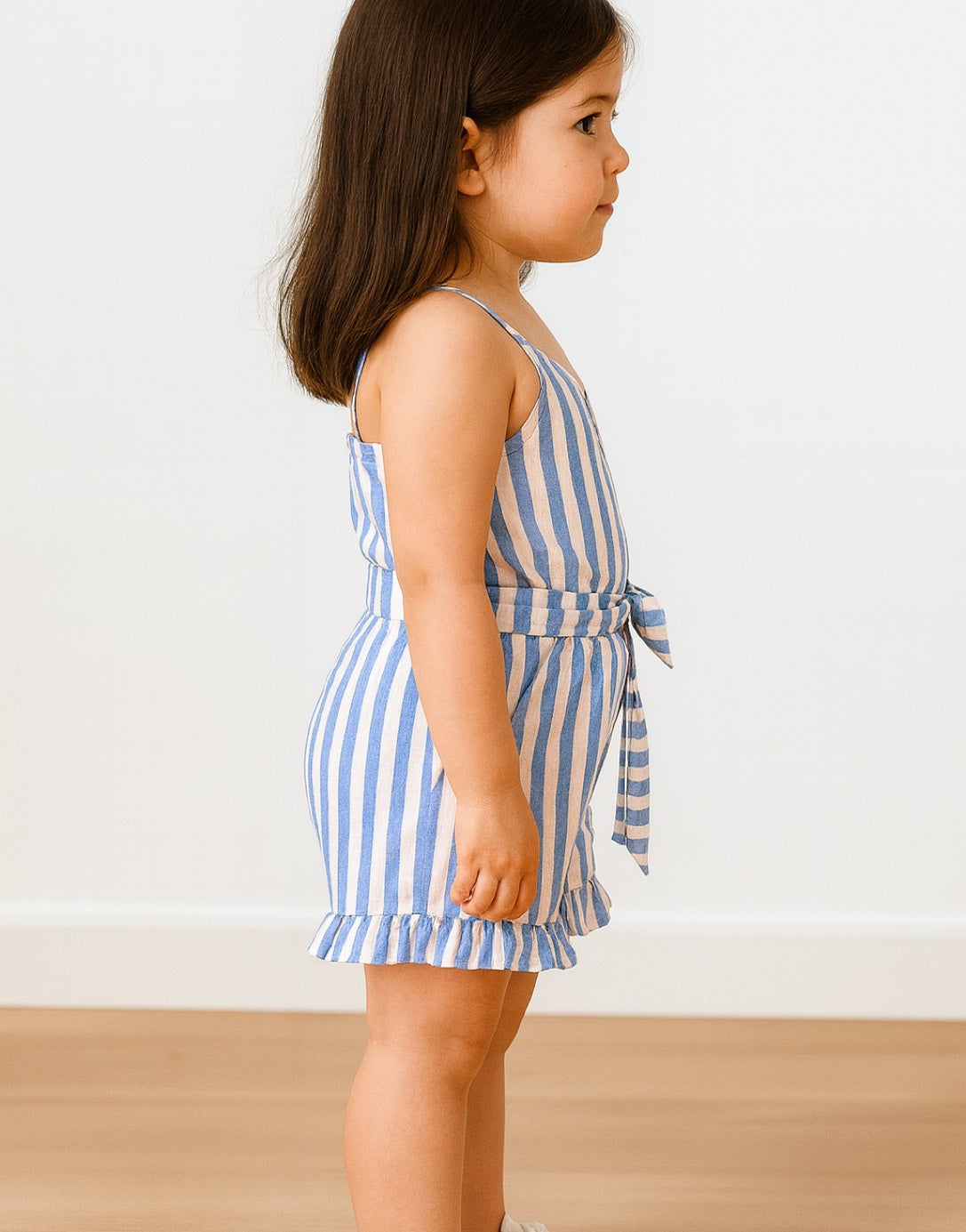 Side view of child wearing a blue and white striped Ruffle Hem Belted Cami Romper against a plain background