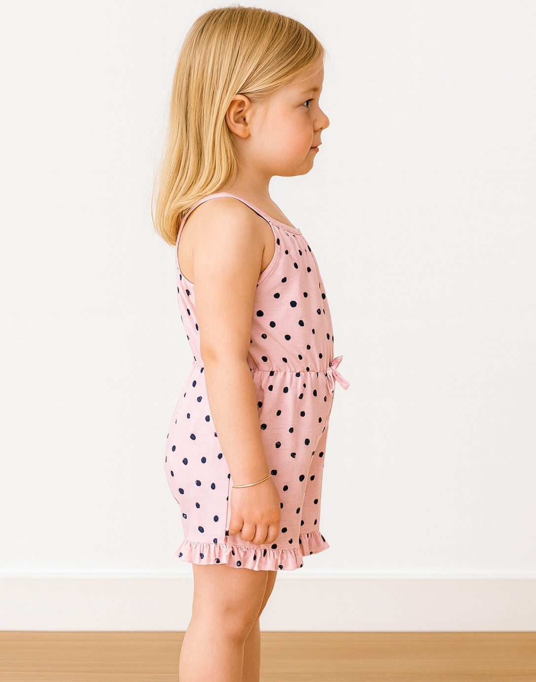 Side view of child wearing a pink polka dot Ruffle Hem Cami Romper on a light wooden floor.