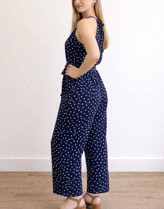 Side view of woman wearing a navy blue Halter Belted Jumpsuit with white polka dots against a plain background.