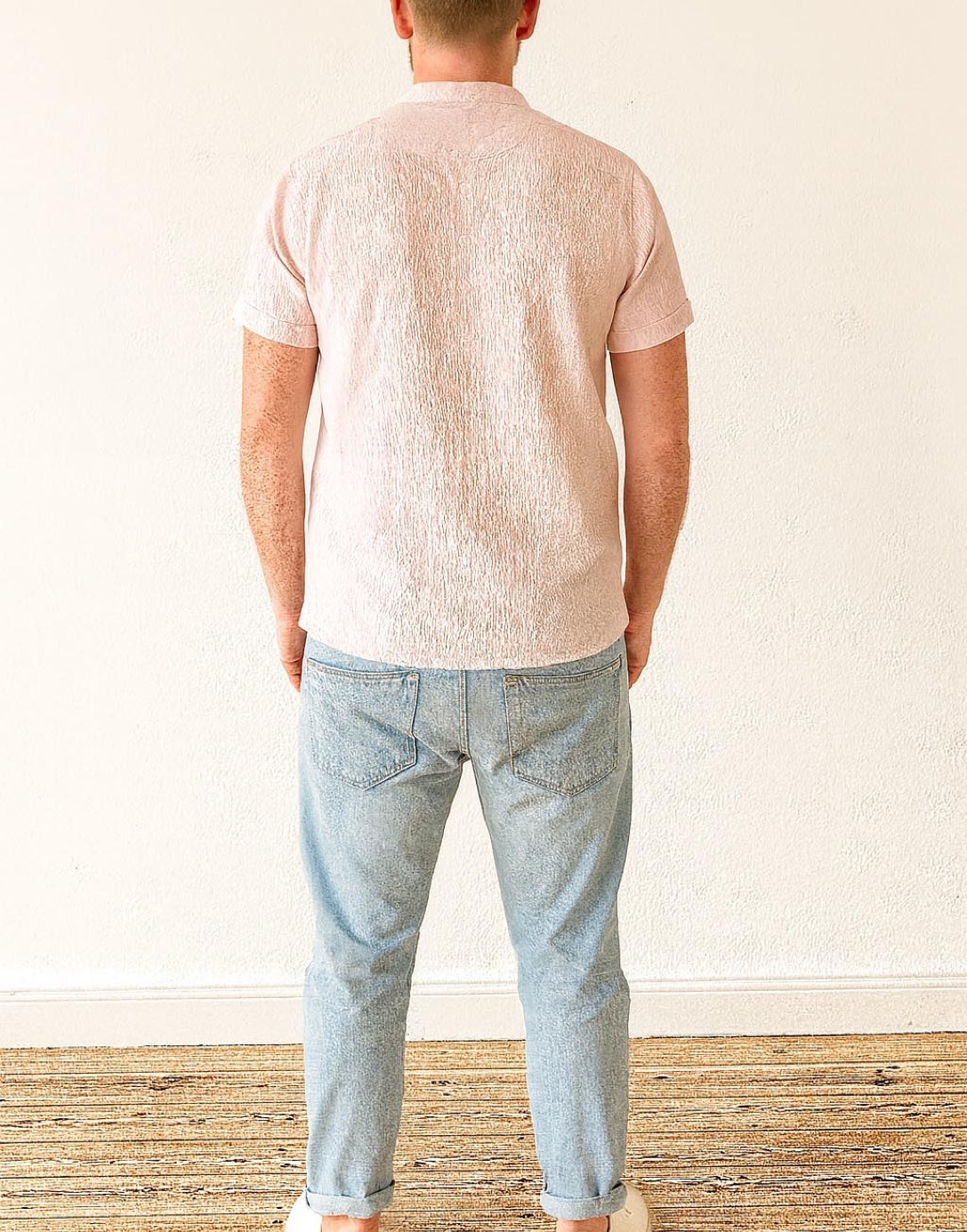Back view of person wearing a light pink Mandarin Collar Short Sleeve Tee Shirt and light blue jeans standing against a white wall.