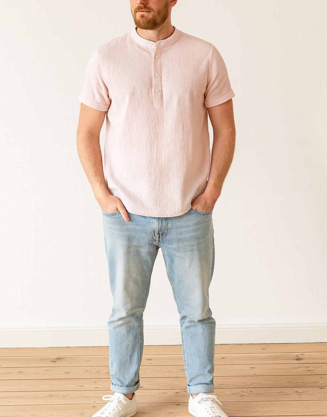Man wearing a light pink Mandarin Collar Short Sleeve Shirt and light blue jeans standing against a white wall.