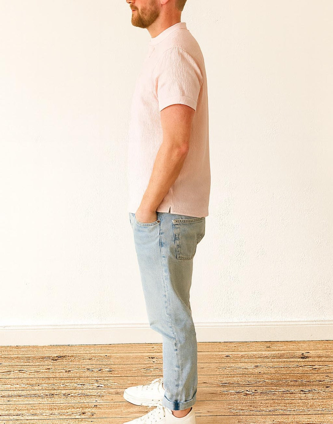 Side view of man wearing a light pink Mandarin Collar Short Sleeve T-Shirt and light blue jeans standing on a wooden floor.