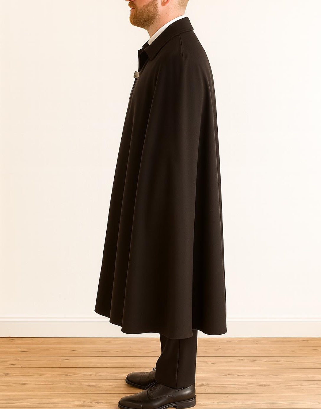 Side view of man wearing a long black Vampire Cape standing on a wooden floor with a white wall background