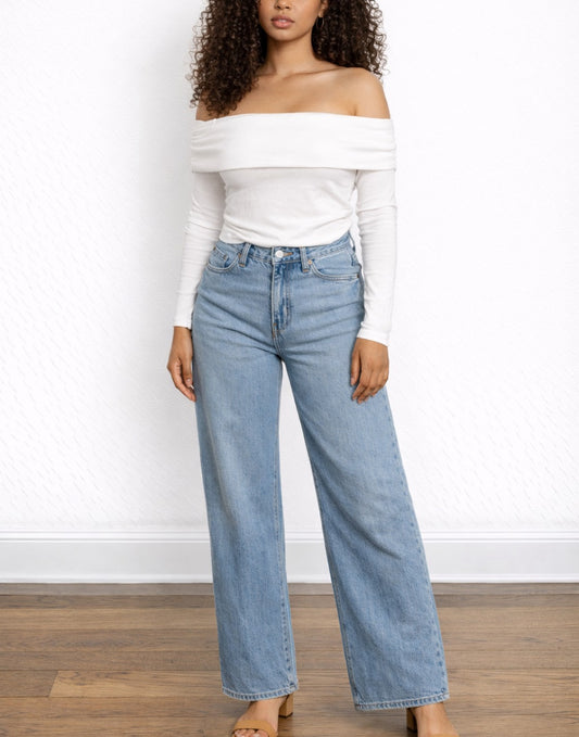 Woman wearing a white Off Shoulder Tee and blue jeans against a white wall.
