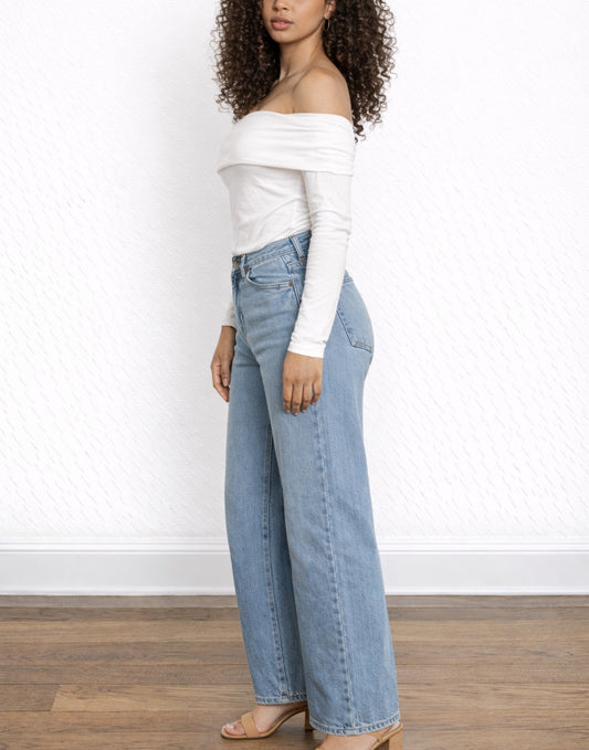 Side view of woman wearing a white Off Shoulder Tee and blue jeans against a white wall.