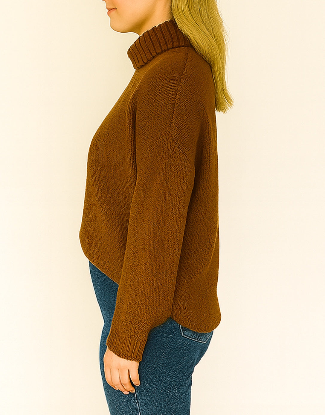 Person turned to the side wearing brown Oversized Turtleneck Sweater and blue jeans standing in front of a plain background.