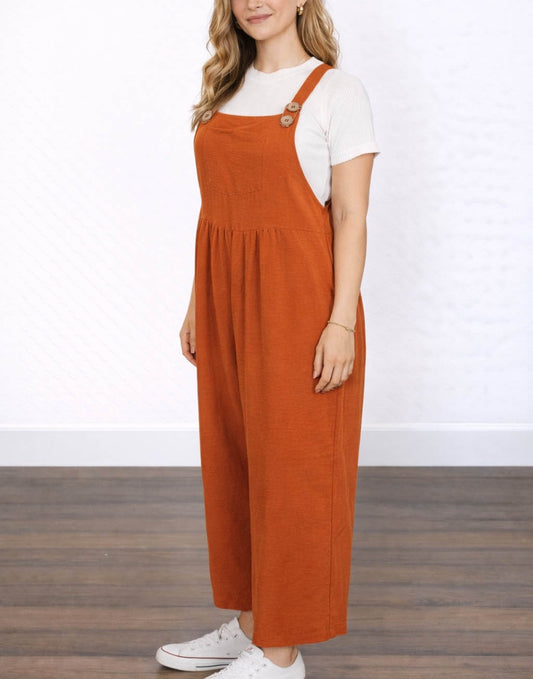 Side view of woman wearing orange Pinafore Jumpsuit with a white shirt underneath on a plain background.