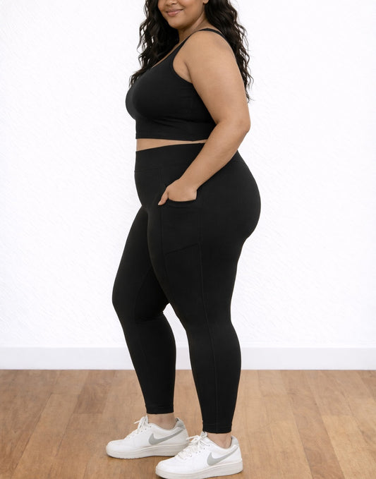 Side view of woman wearing a black athletic top and Plus Size Side Pocket Leggings with white sneakers on a wooden floor.