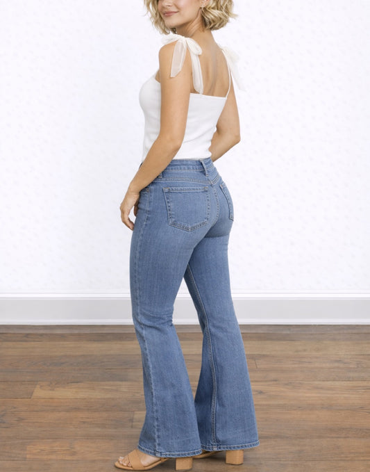 Side view of woman wearing a white Shoulder Tie Tank Top and blue jeans standing on a wooden floor with a white wall background.