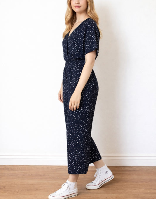 Side view of woman wearing a navy blue Surplice Jumpsuit with white polka dots on a plain background.