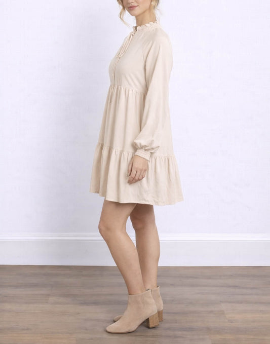 Side view of woman wearing a beige Tie Neck Smock Mini Dress with long sleeves against a white background.