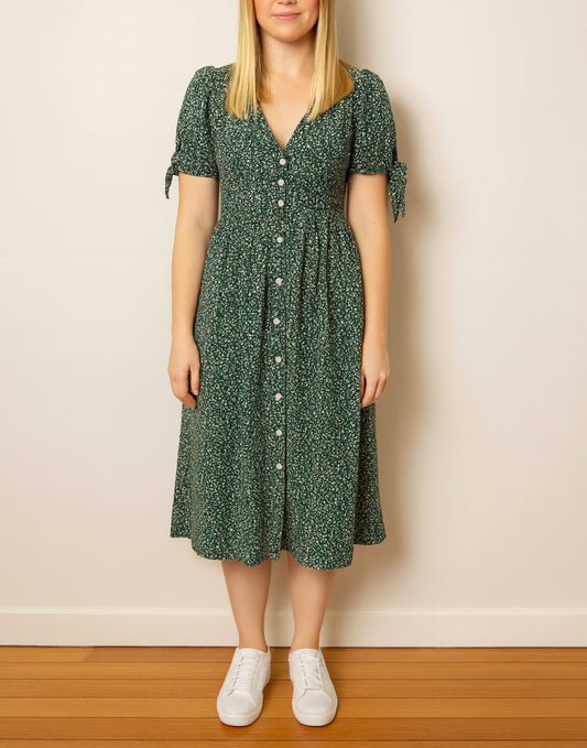 Woman wearing a green floral Tie Sleeve Button Front Midi Dress against a plain wall.