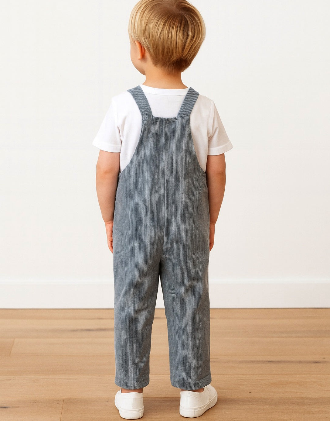 Back view of child wearing blue overalls standing on a wooden floor with a white background