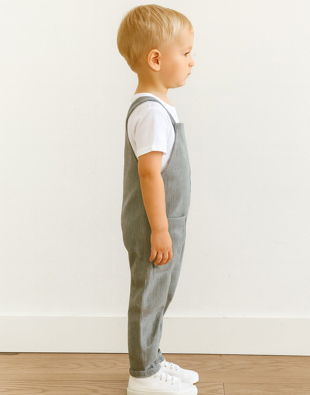 Side view of child wearing gray overalls standing against a white wall.