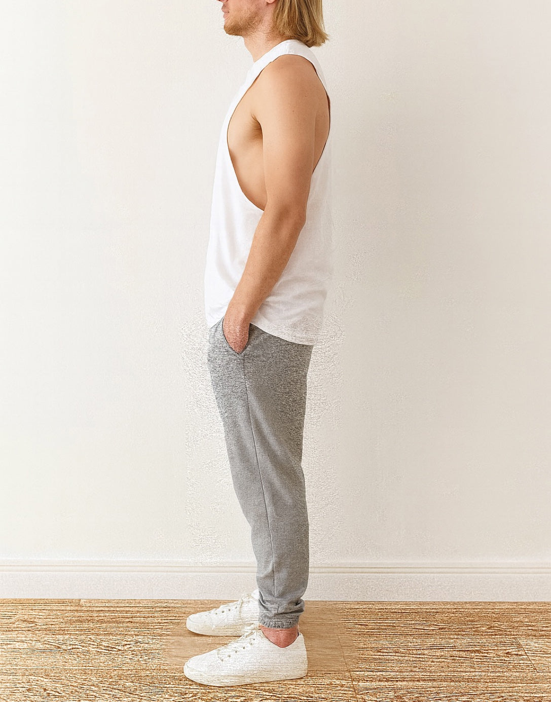 Side view of person wearing a white Workout Tank Top and gray sweatpants standing against a plain wall.