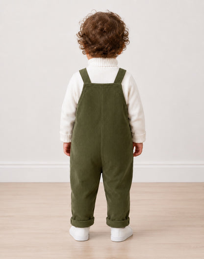 Back view of a toddler standing indoors wearing olive green corduroy overall-style pants over a white ribbed turtleneck sweater, with white sneakers, against a plain light wall and wood floor.